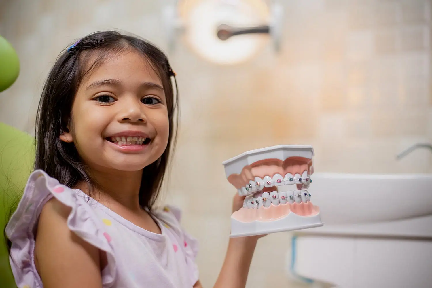 At Tollison Orthodontics in Greenville, SC, a young girl smiles and holds a dental model with braces while sitting in the children's orthodontist clinic.