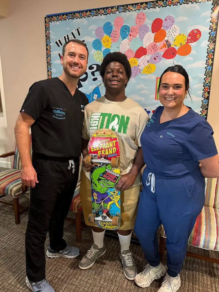 At Tollison Orthodontics in Greenville, SC, three smiling people pose indoors, with the center person holding a colorful skateboard; a bulletin board covered in heart-shaped notes appears behind them.