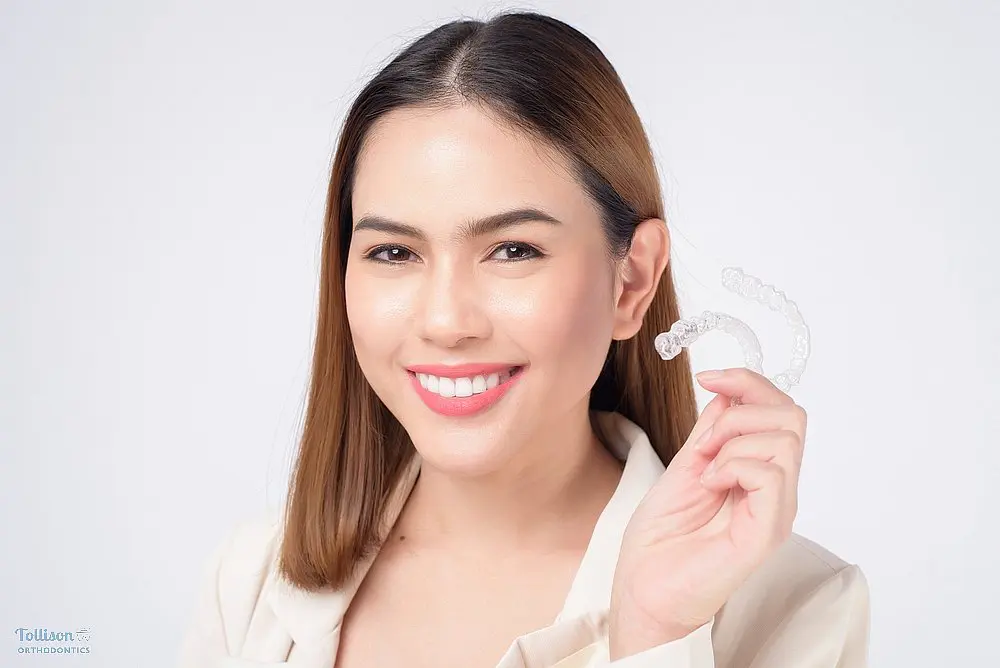 Smiling young woman holding an Invisalign clear aligner as one option of alternatives to braces in Greenville, SC in her hand against a white background.
