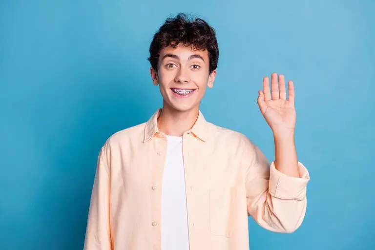 A teenager with curly hair, braces, and a light peach shirt over a white t-shirt stands smiling and waving in front of a blue background at Tollison Orthodontics in Greenville, SC. Wondering how long do braces take in Greenville?.