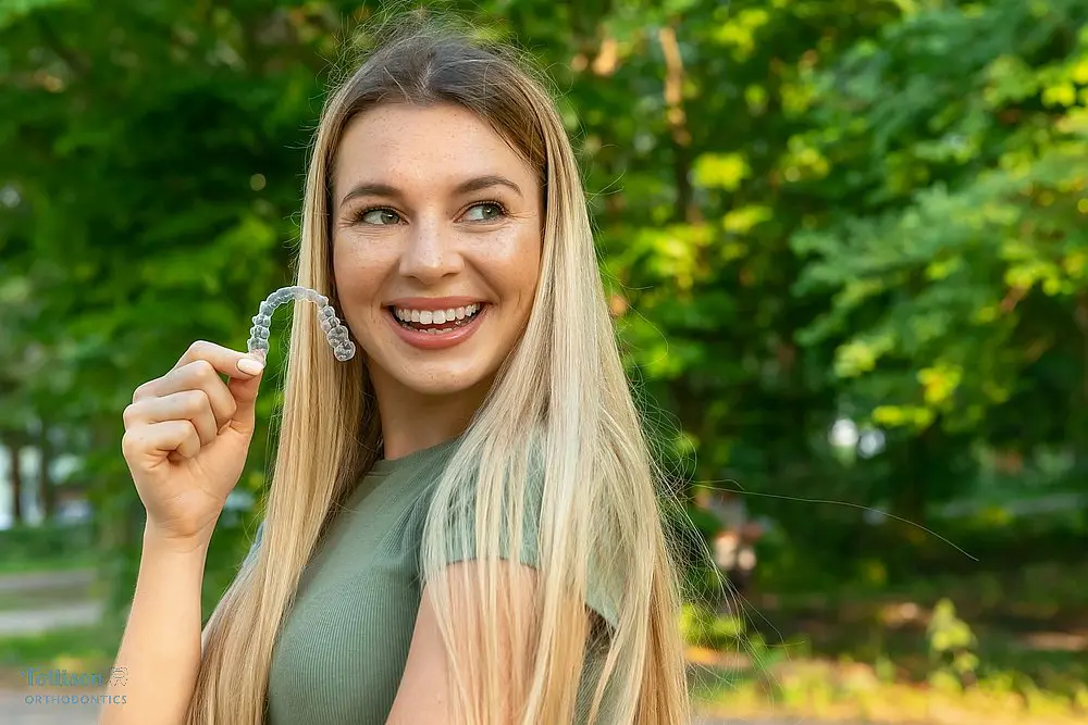 Young woman with long blonde hair smiling while holding a clear aligner outdoors after understanding Does Insurance Cover Invisalign in Greenville, SC, showcasing Invisalign treatment.