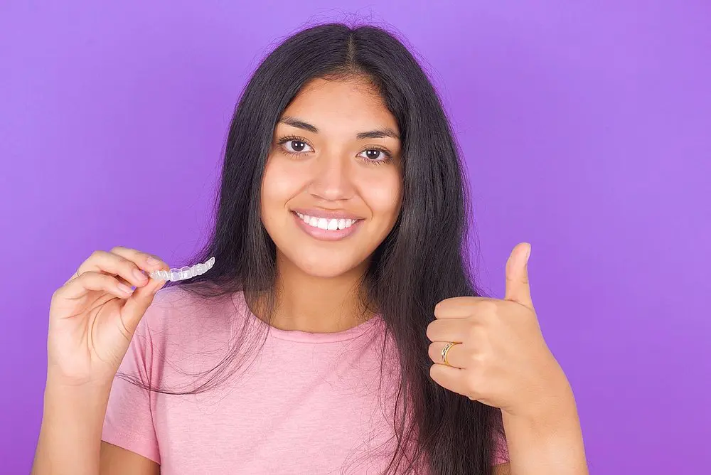 A teenager with curly hair, braces, and a light peach shirt over a white t-shirt stands smiling and waving in front of a blue background at Tollison Orthodontics in Greenville, SC. Wondering how long do braces take in Greenville?.