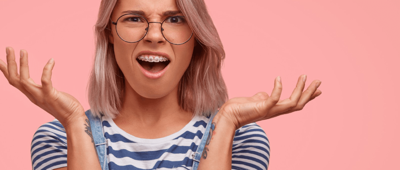 A person with glasses and adult orthodontics is wearing a striped shirt, raising their hands in surprise against a pink background in Greenville, SC