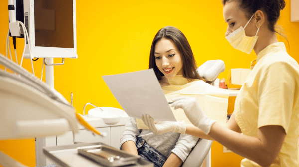 A dentist wearing a mask holds a document detailing orthodontic insurance plans while a patient sits in the dental chair, smiling. Surrounding them is an array of dental equipment, all set against the backdrop of an orange wall in Greenville, SC
