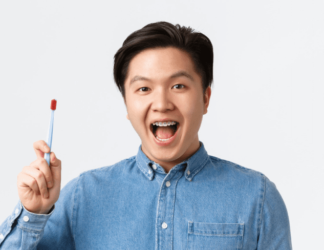 A person in a denim shirt holds a toothbrush and smiles with mouth open, proudly showcasing the results of adult orthodontics against a plain background in Greenville, SC