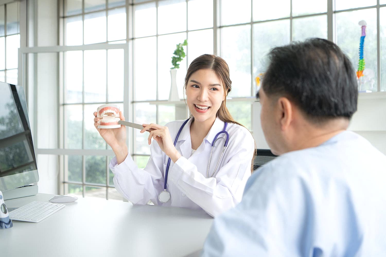 A dentist with a stethoscope discusses adult braces while showing a dental model to a patient in an office with large windows in Greenville, SC