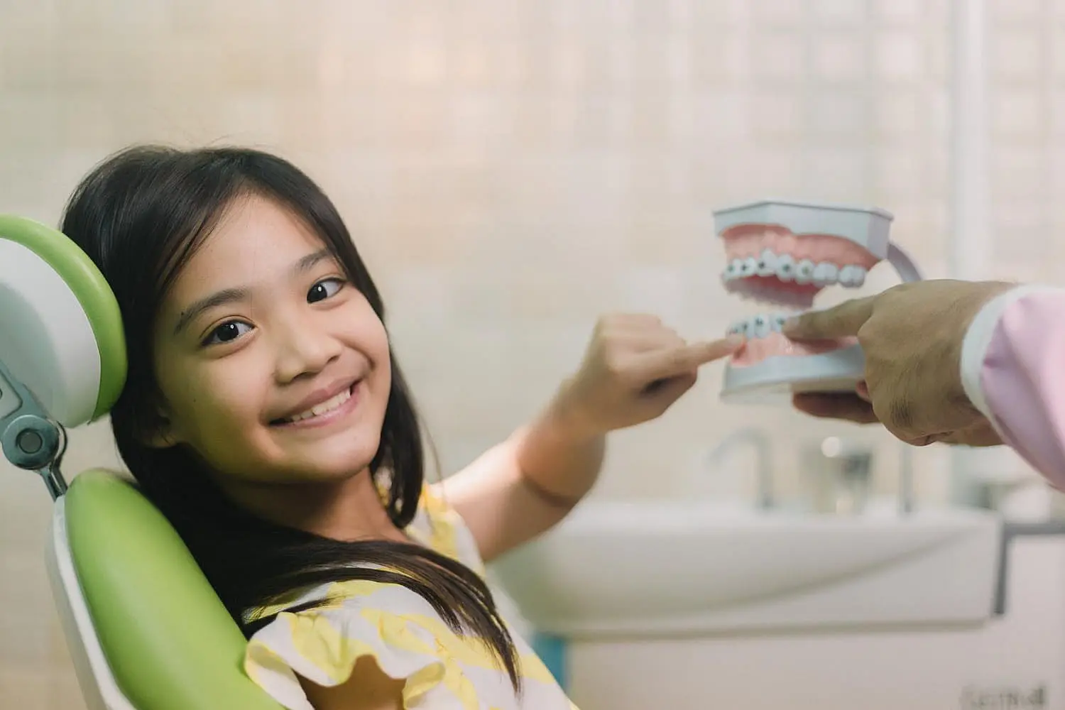 A young girl in a dental chair smiles while pointing at a dental model held by her friendly children's orthodontist in Greenville, SC