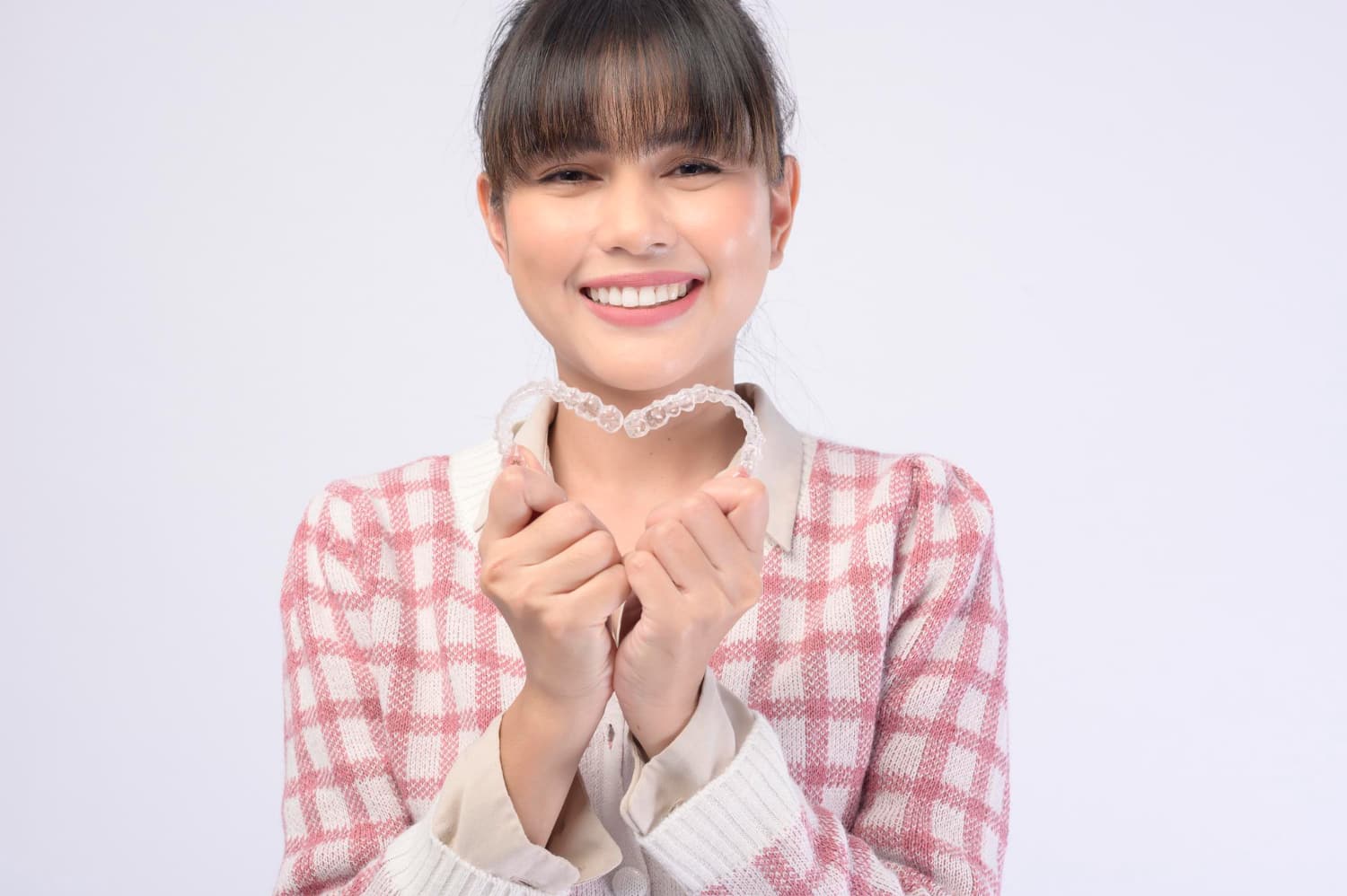 A person smiling, holding an Invisalign clear dental aligner with both hands in front of a plain background, showcasing the benefits of adult orthodontics while wearing a pink and white checkered sweater in Greenville, SC