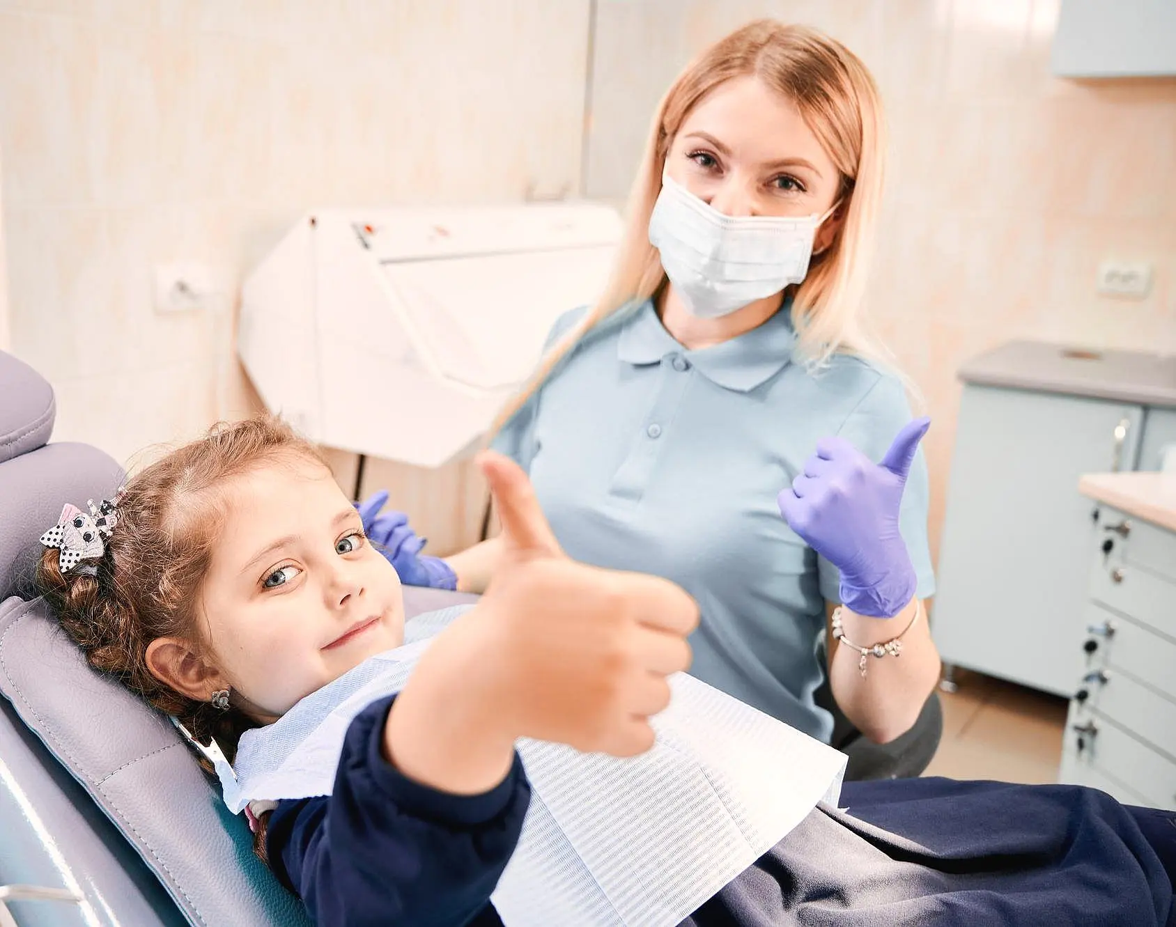 A children's orthodontist wearing a mask and gloves gives a thumbs up alongside a young girl in the dental chair, both beaming positivity in the dental office setting in Greenville, SC