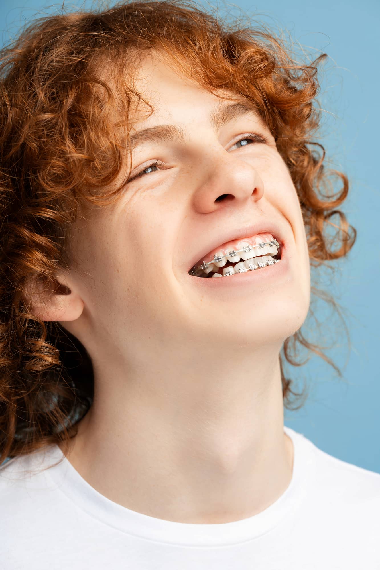 A person with curly red hair grins confidently, showcasing their braces—an example of successful adult orthodontics—while wearing a white shirt against a vibrant blue background in Greenville, SC