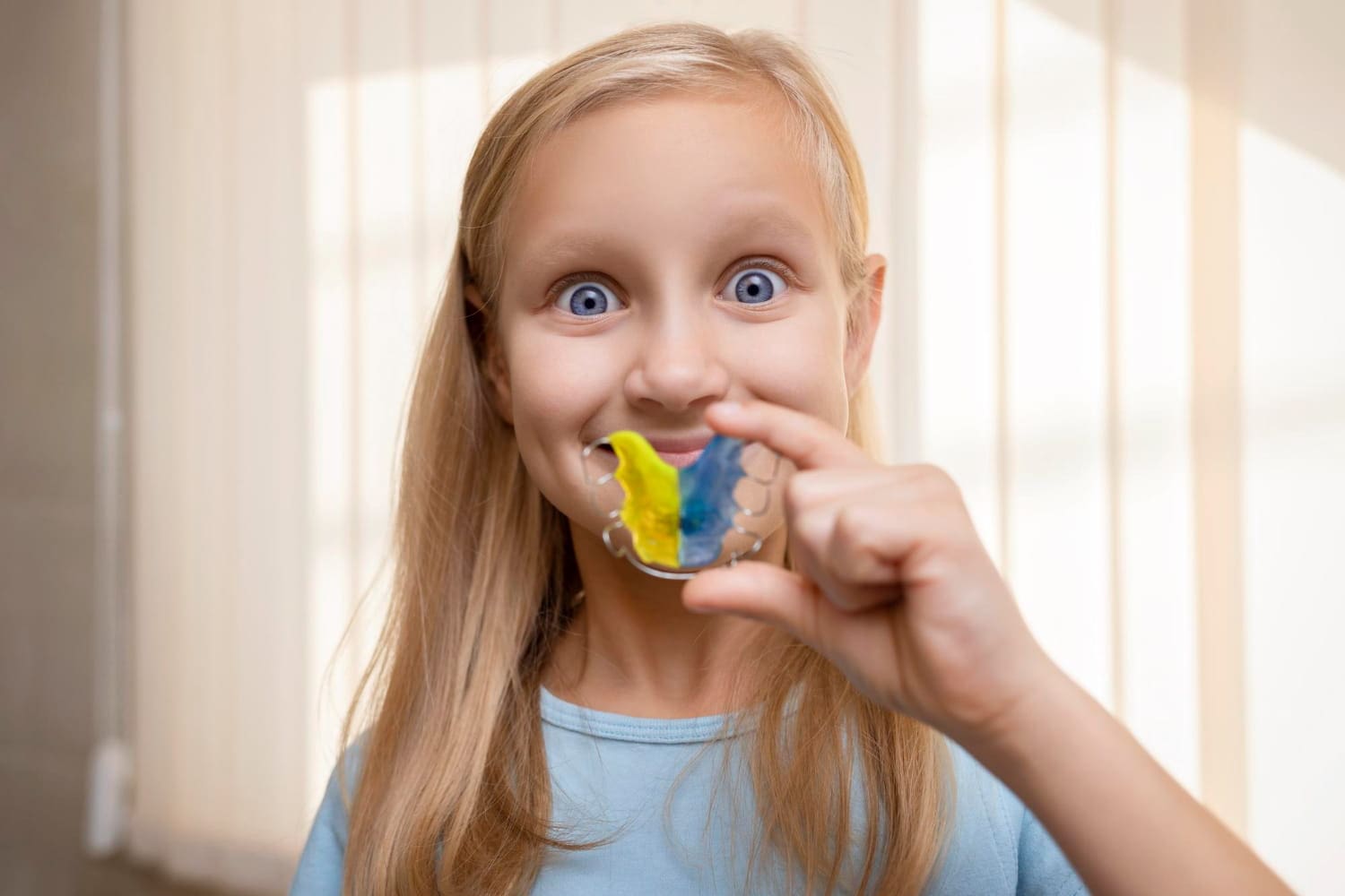 A girl with long hair holds a colorful dental retainer in front of her mouth, smiling widely, perhaps imagining her next visit to the children's orthodontist. She wears a light blue shirt, with vertical blinds providing a subtle backdrop in Greenville, SC