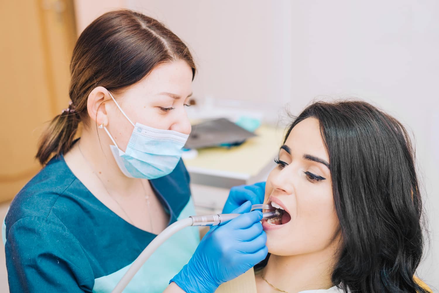 A dentist, equipped with orthodontic appliances, wears a mask and blue gloves as they use dental tools to meticulously examine a woman's teeth in the dental office in Greenville, SC