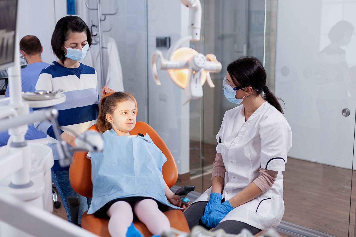 A young girl sits in a dental chair wearing a bib, accompanied by an adult. A children’s orthodontist in a white coat and mask discusses treatment options. Dental equipment is visible in the room in Greenville, SC