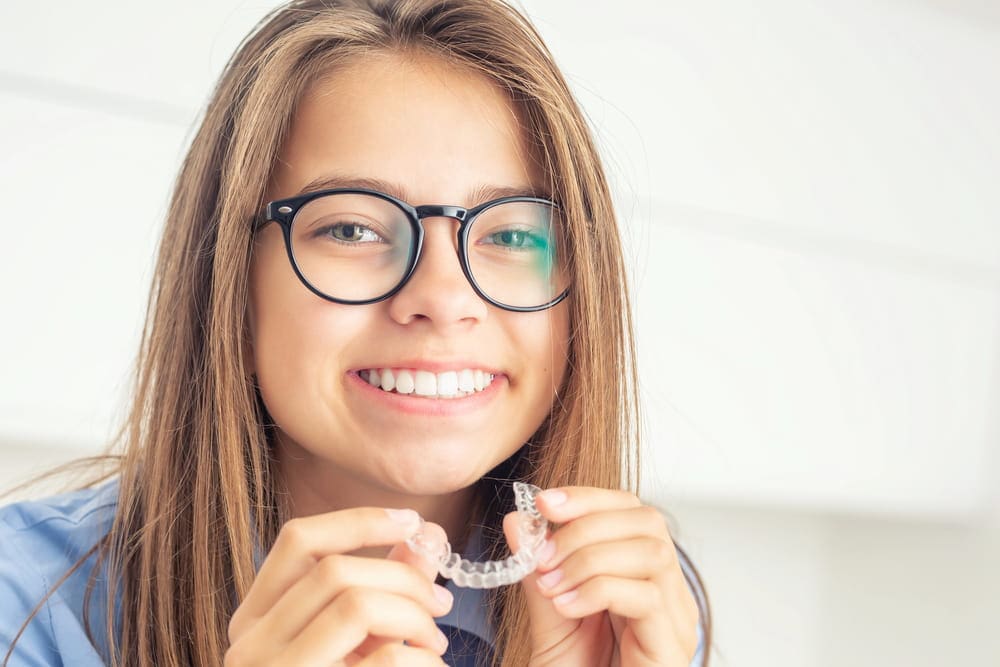 A person with glasses smiles while holding a clear dental retainer, as if just returning from a visit to their children's orthodontist in Greenville, SC