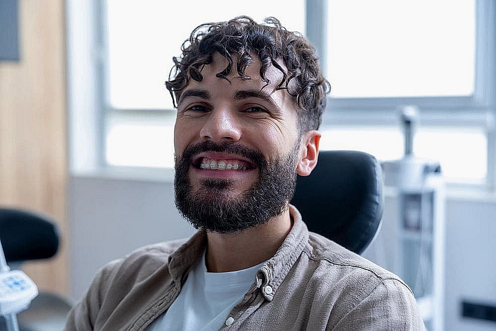 A man with curly hair and a beard smiles while seated indoors, wearing ceramic braces, a brown shirt, and a white t-shirt. The background features a window and blurred office furniture in Greenville, SC