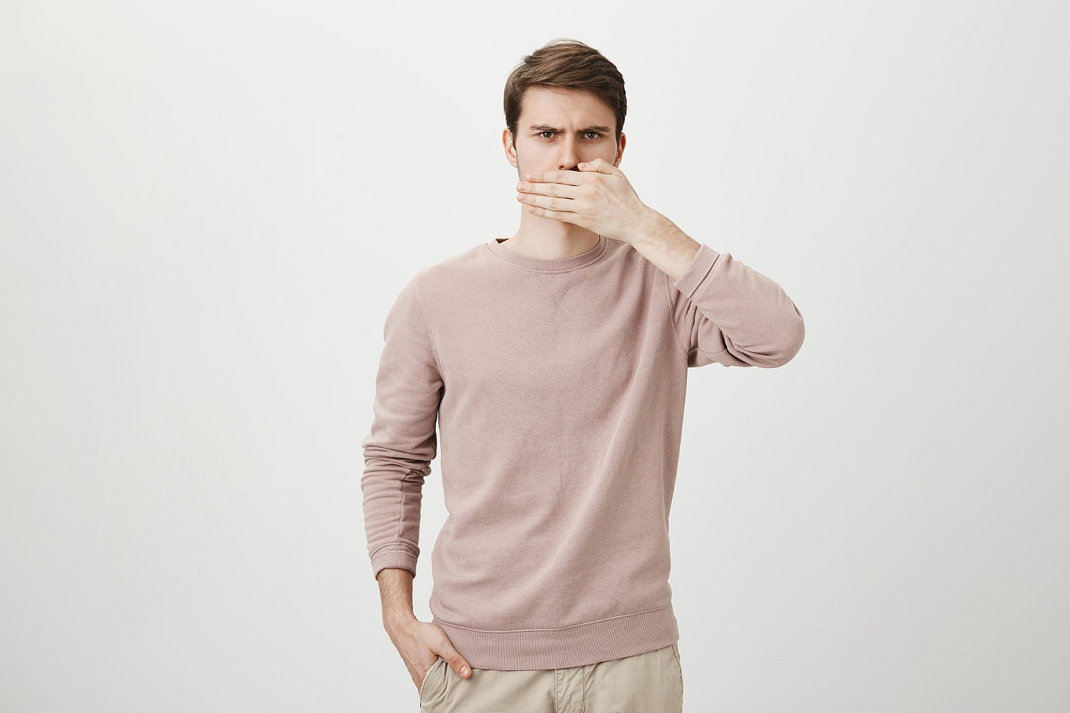 A man in a pink sweater covers his mouth with his hand, eyeing the camera with a knowing look, reminiscent of a thoughtful yet silent mouth breather against a plain background in Greenville, SC