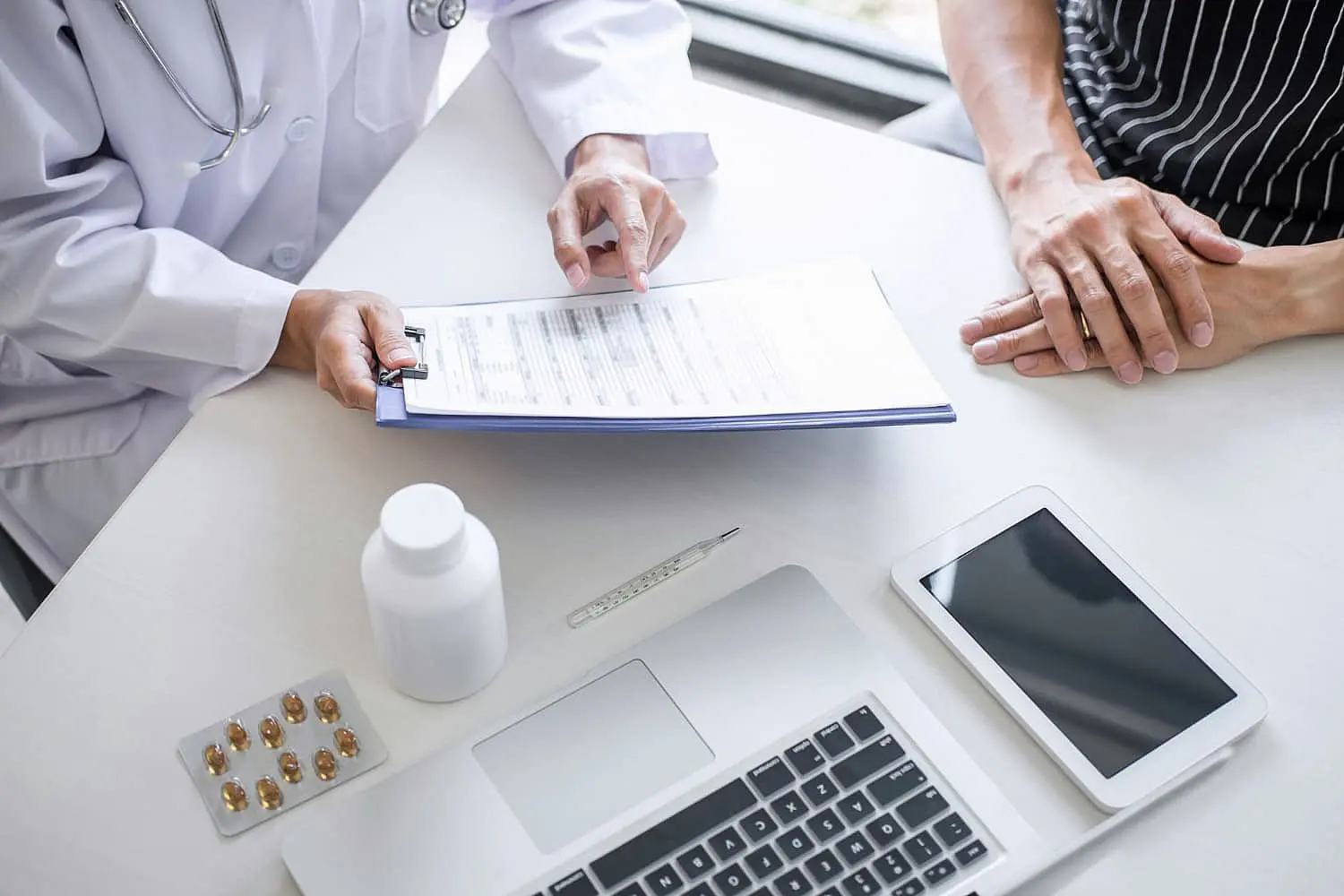 A doctor discusses orthodontic insurance plans with a patient, showing a clipboard with documents across a desk adorned with a laptop, tablet, pills, and a bottle in Greenville, SC