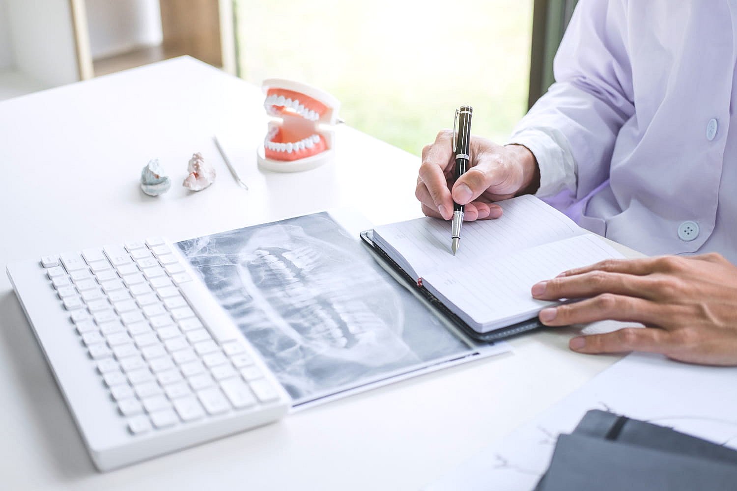 A person in a lab coat diligently writes in a notebook at a desk, surrounded by dental molds, X-ray images, and a keyboard, meticulously reviewing orthodontic insurance plans in Greenville, SC