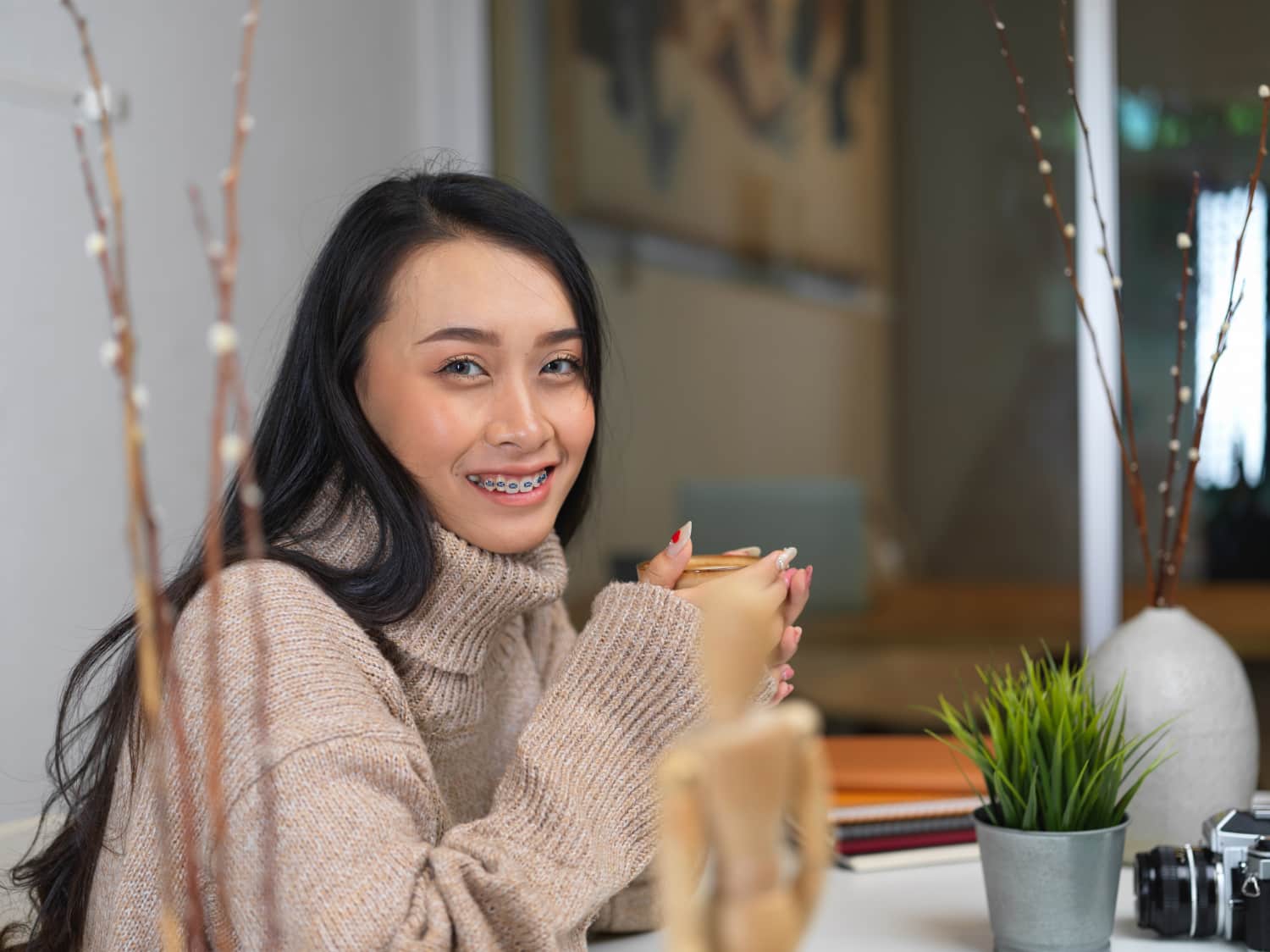 A person with long hair and braces, showcasing their journey in adult orthodontics, smiles while holding a mug, seated at a table adorned with plants, books, and a camera in Greenville, SC