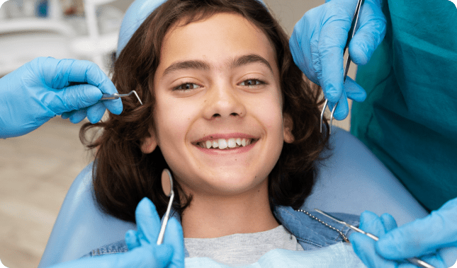 A smiling child at the orthodontist is surrounded by hands in blue gloves holding dental tools in Greenville, SC