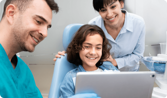 A smiling child sits happily in a dental chair, looking at a tablet held by the orthodontist. Nearby, the dental assistant watches with a warm smile, ensuring a comforting experience for their young patient in Greenville, SC