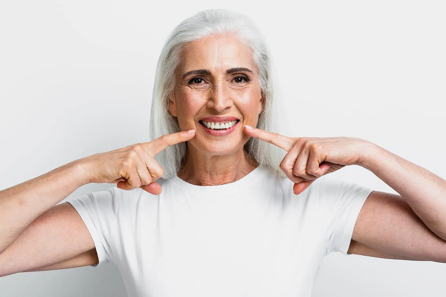 An older woman with long white hair, clad in a white shirt, smiles at the camera while pointing confidently to her teeth with both index fingers against a plain white background, showcasing the results of her orthodontic insurance plans in Greenville, SC