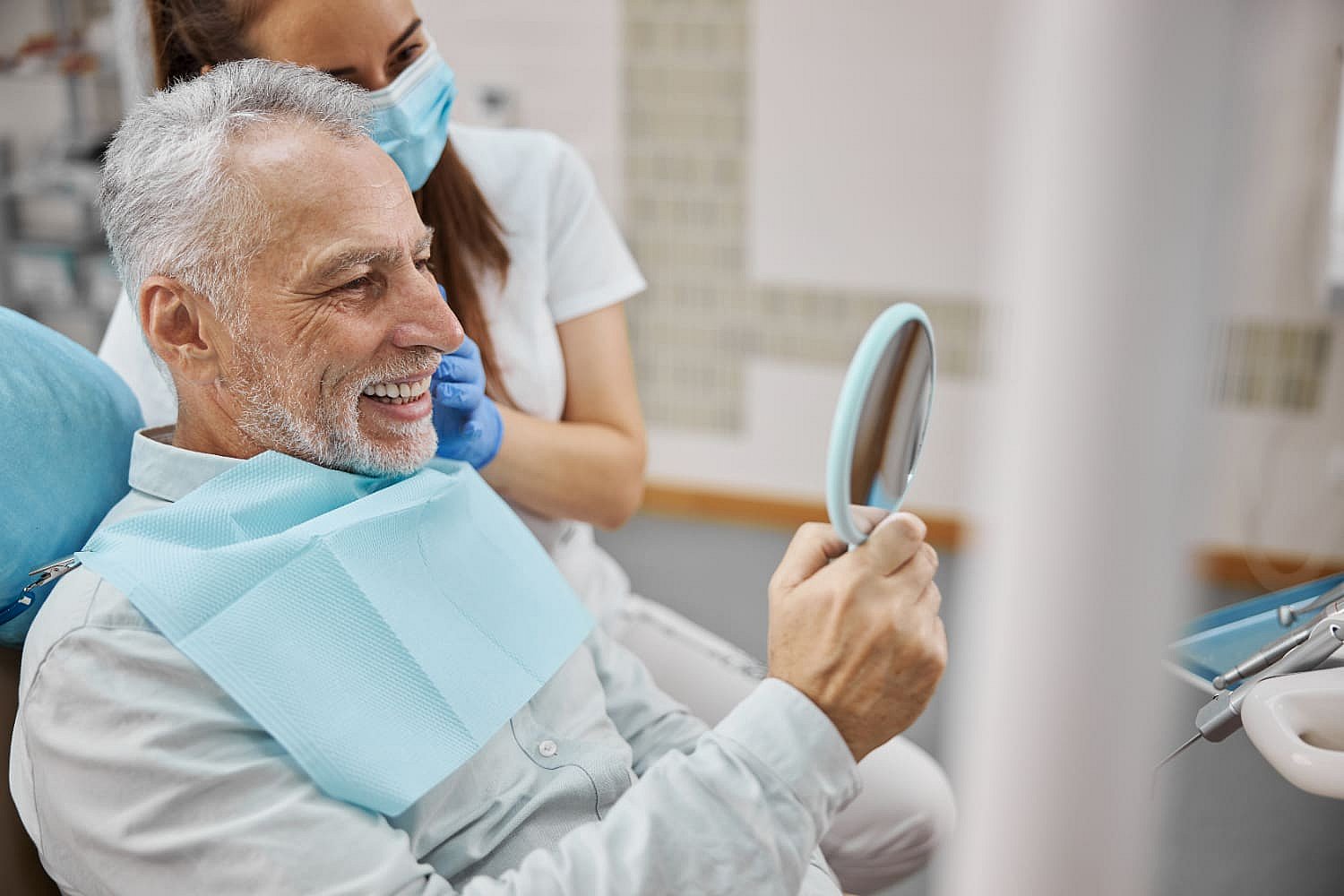 An older man smiles confidently in a dental chair, holding a mirror, as a female dental professional in a mask and gloves stands beside him, showcasing the remarkable results of adult orthodontics in Greenville, SC