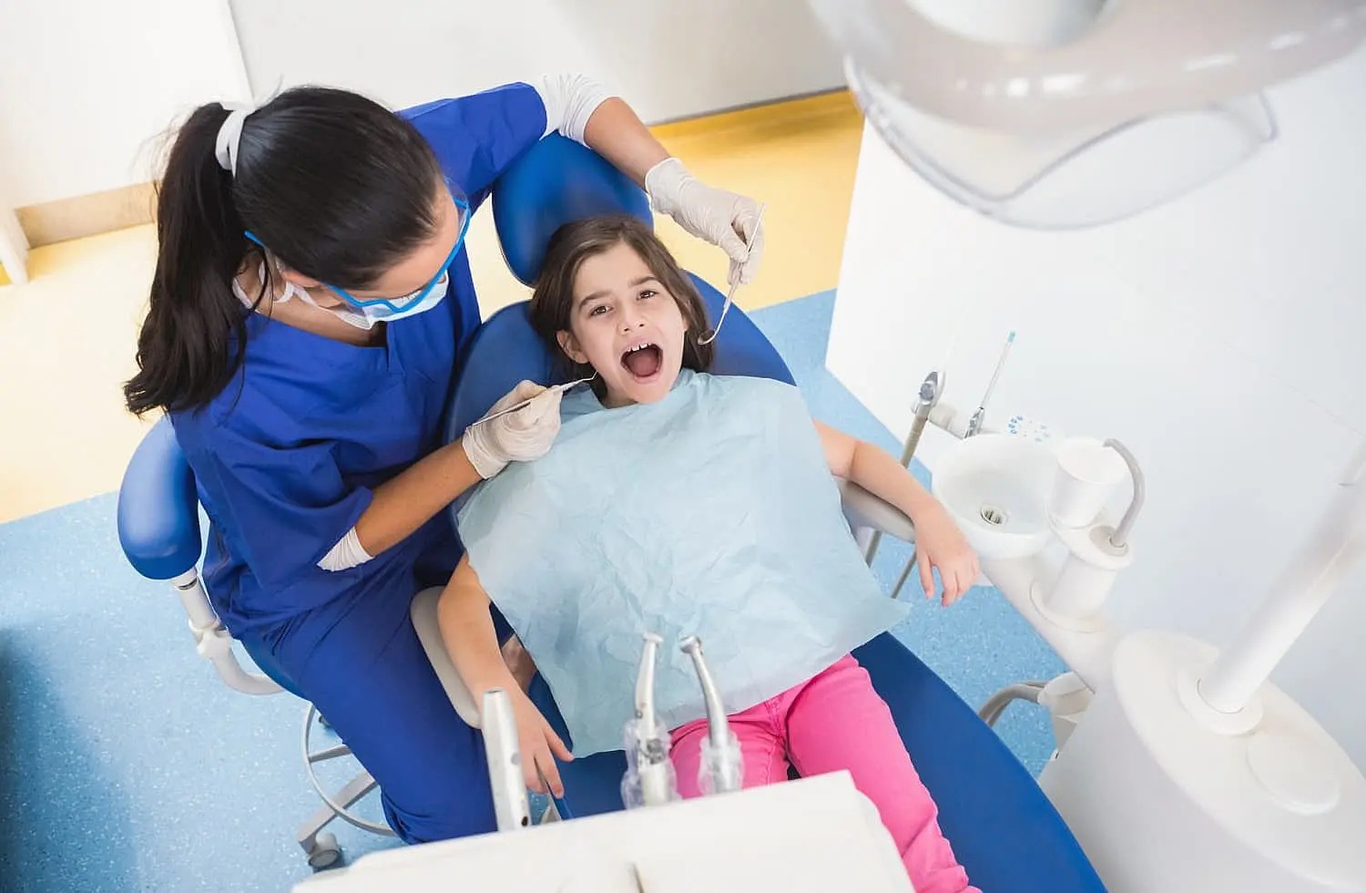 A children's orthodontist examines a child in a dental chair. The young patient wears a bib and has her mouth open, ready for the check-up. The orthodontist is dressed in blue scrubs and gloves, skillfully using dental tools to ensure everything is in order in Greenville, SC