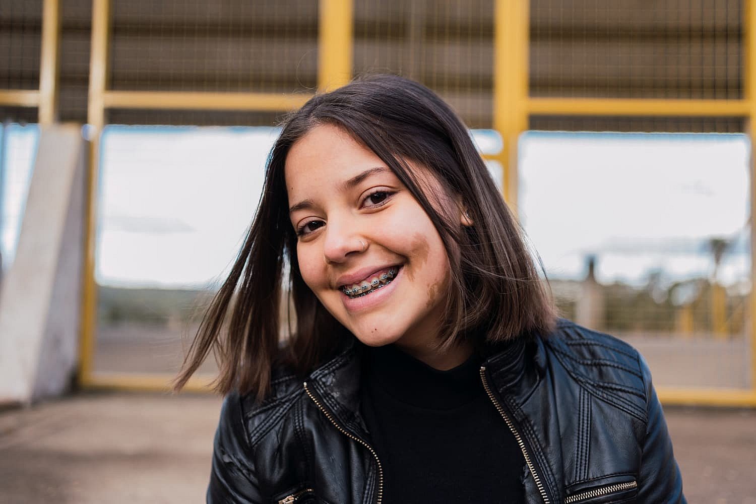 A young woman with shoulder-length hair and a radiant smile, her braces gleaming, stands in a black jacket outdoors with yellow metal fencing in the background in Greenville, SC