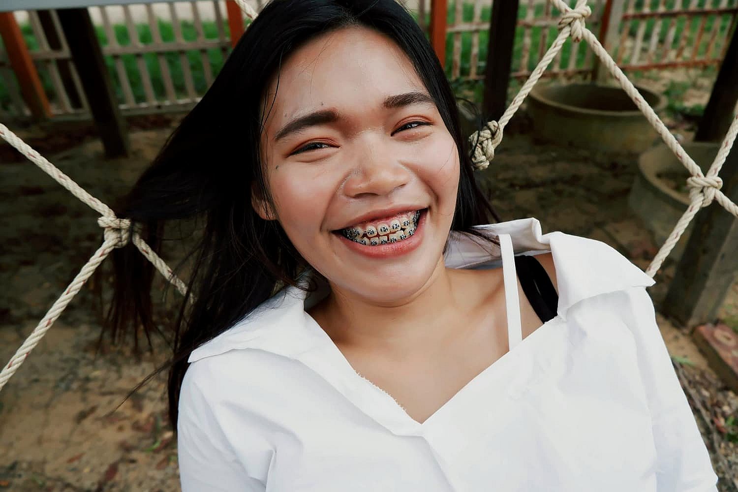 A person with long hair and braces, enjoying the benefits of orthodontic insurance plans, smiles broadly while wearing a white shirt and leaning against a rope net outdoors in Greenville, SC
