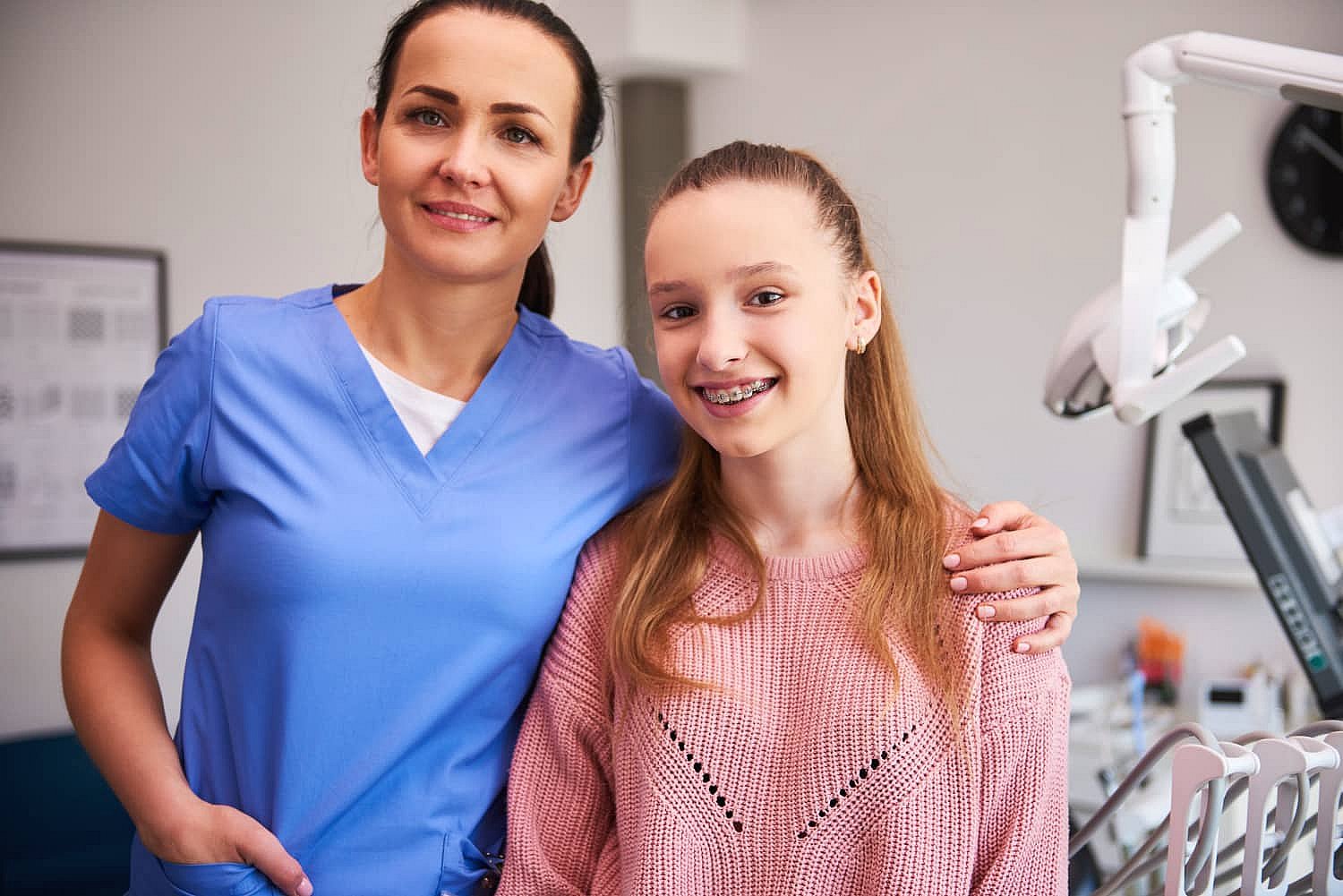 A dental professional in blue scrubs stands smiling with a young girl proudly wearing metal braces in a dental office in Greenville, SC
