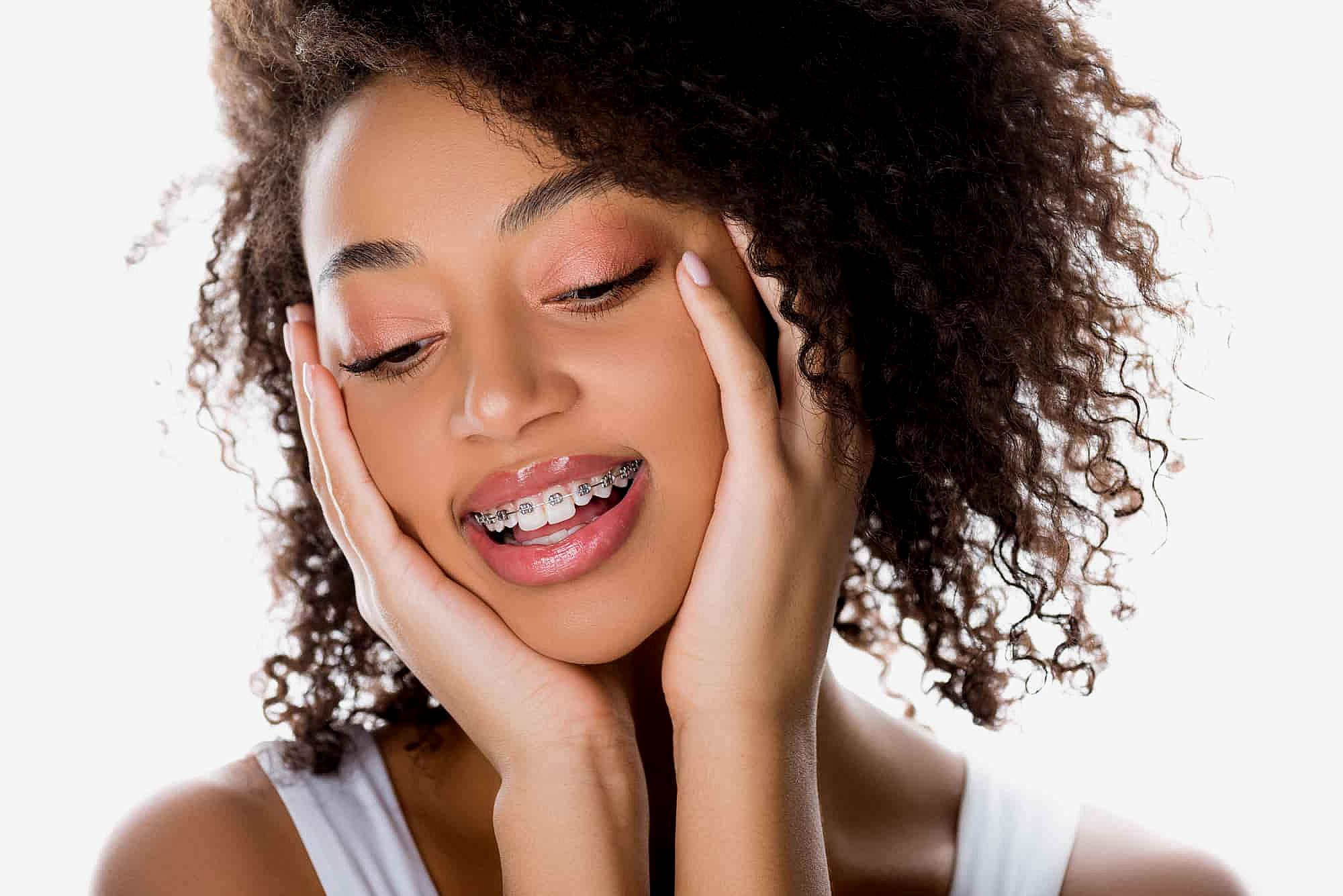 A person with curly hair and braces smiles, showcasing their unique style of orthodontics while gently holding their face with both hands in Greenville, SC