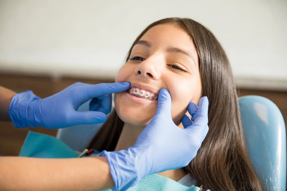 A children's orthodontist with blue gloves examines a girl's braces as she sits in a dental chair, smiling slightly in Greenville, SC