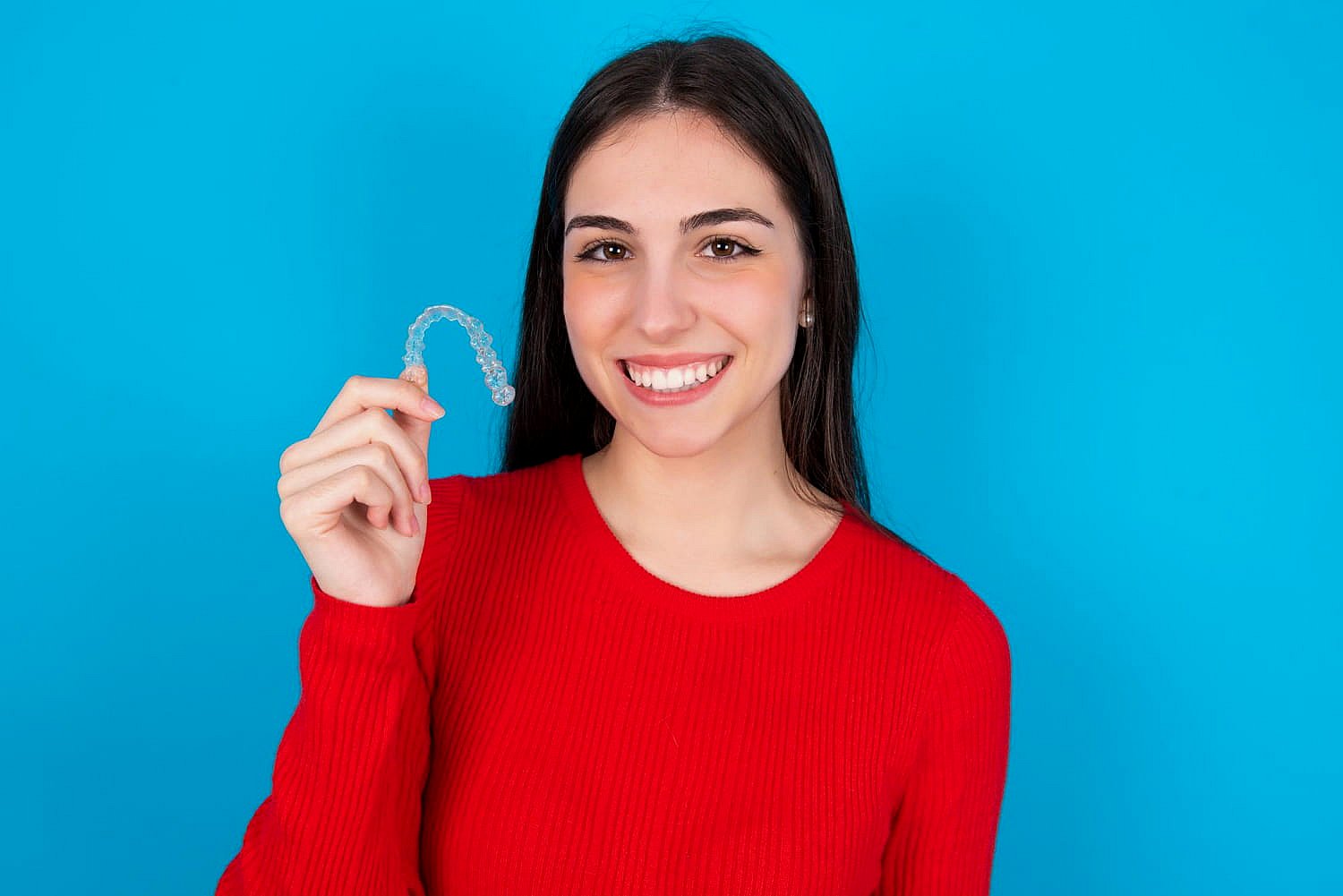 A smiling individual in a red sweater proudly displays their Invisalign clear dental aligner against a vibrant blue background in Greenville, SC