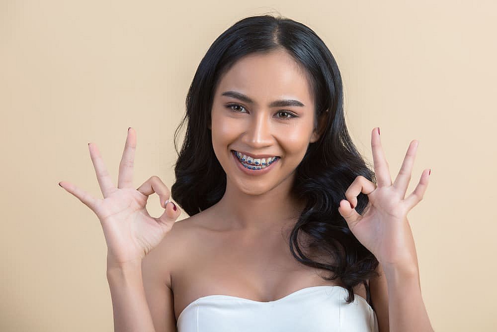 A woman with long black hair and metal braces smiles, making "OK" hand gestures against a plain beige background. She wears a strapless white top in Greenville, SC