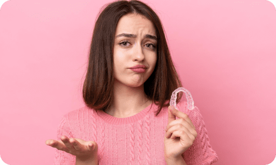 A woman in a pink sweater holds a dental retainer in one hand and gestures with the other against a pink background, her slightly puzzled expression suggesting she's contemplating orthodontic insurance plans in Greenville, SC