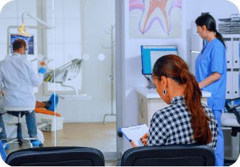 A woman fills out a form in a dental office waiting area, pondering orthodontic insurance plans. In the background, a dentist examines a patient while a nurse taps away on a computer in Greenville, SC