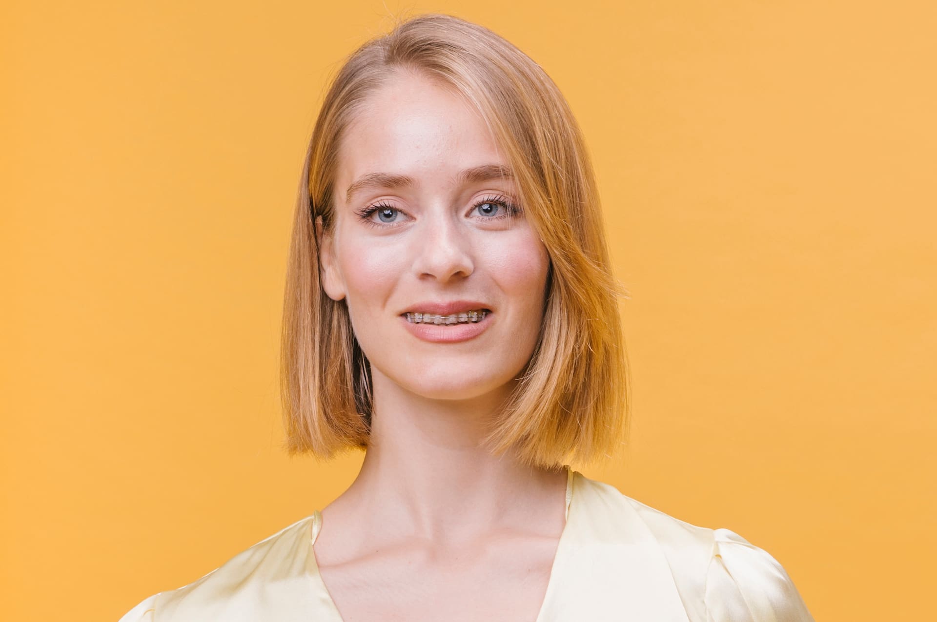 A woman with shoulder-length blonde hair and clear braces smiles in front of a solid yellow background. Embracing adult orthodontics, she confidently wears a light yellow top in Greenville, SC