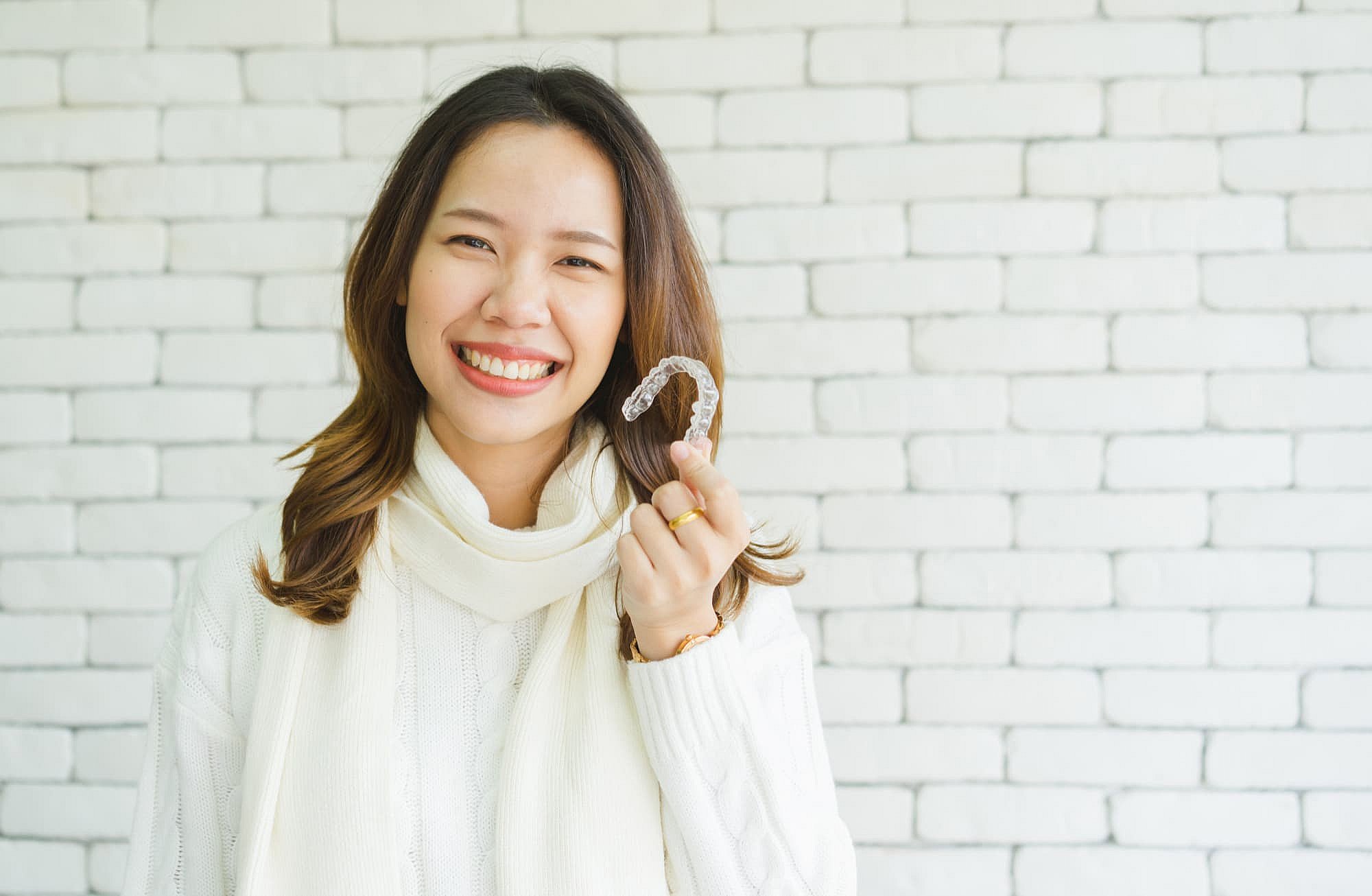 A woman beams brightly, showcasing her Invisalign clear aligner in front of a white brick wall. Dressed in a cozy white sweater and scarf, her smile captures the transformative power of modern dental care in Greenville, SC