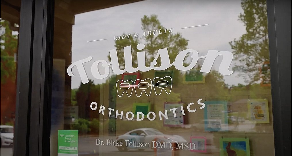 A glass door at Tollison Orthodontics in Greenville, SC features three tooth illustrations, Dr. Blake Tollison's name, and colorful flyers.