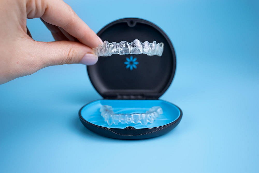 A hand holds a transparent dental invisalign clear aligner above an open black and blue case containing another aligner, set against a solid blue background at Tollison Orthodontics Greenville SC